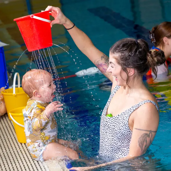 Parent holds a baby at water level in an indoor pool during a WaterBabies lesson, with a swim teacher guiding the activity nearby.