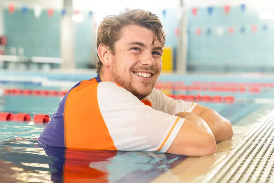 Adult swimmer practises technique in an indoor lap pool while a Carlile Swim instructor coaches from the water beside them.