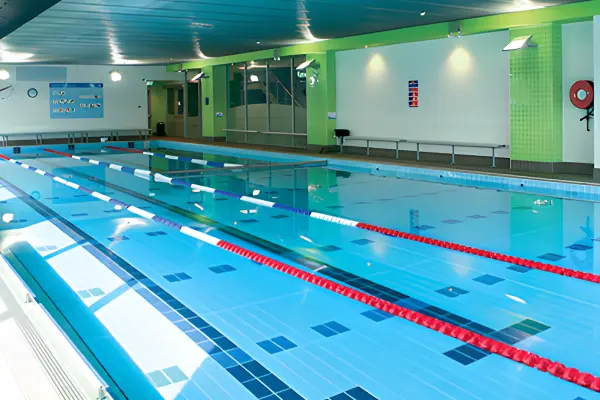 Indoor lap pool at Carlile Swimming Five Dock facility, showing clear blue water, tiled lanes with red and blue lane ropes, and the pool deck with green-and-white walls.