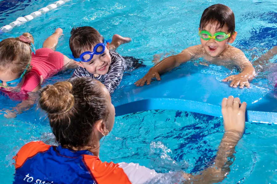 Swim teacher in a blue-and-orange vest coaches three preschool children wearing goggles as they kick and hold a blue foam float in an indoor pool.
