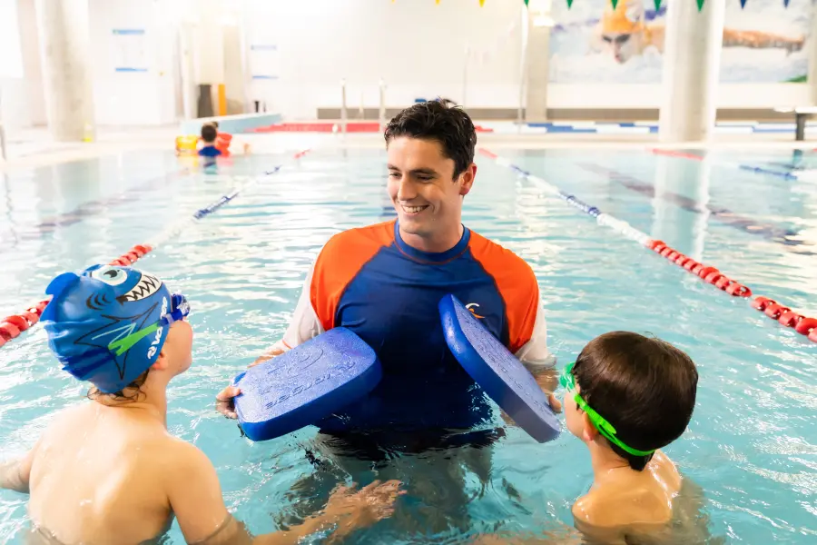 Swim teacher watches and coaches a child swimming in an indoor pool lane during a stroke correction session, focusing on technique and streamlined body position.