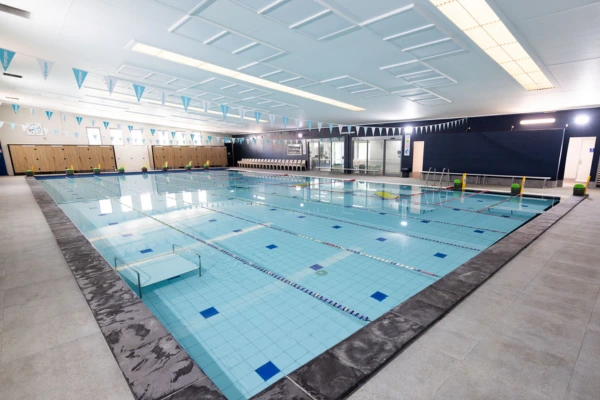 Indoor heated lap pool at Carlile Swim North Lakes, showing marked lanes with lane ropes, tiled pool deck, and the bright indoor swim centre.