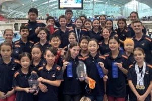 Group of Carlile Swim squad swimmers and coaches posing on the pool deck at a swim meet, wearing navy uniforms and holding medals, ribbons, and trophies.