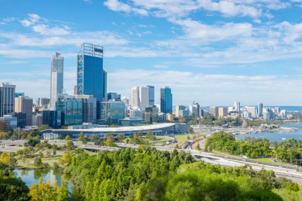 Perth skyline during the day, featuring modern high-rise buildings along the Swan River, with Kings Park greenery and winding highways in the foreground under a bright blue sky.