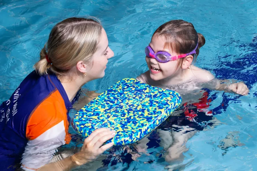 A swim instructor in a blue and orange uniform smiles at a young girl wearing pink and purple goggles during a one-on-one swimming lesson, with a kickboard between them in an indoor pool.