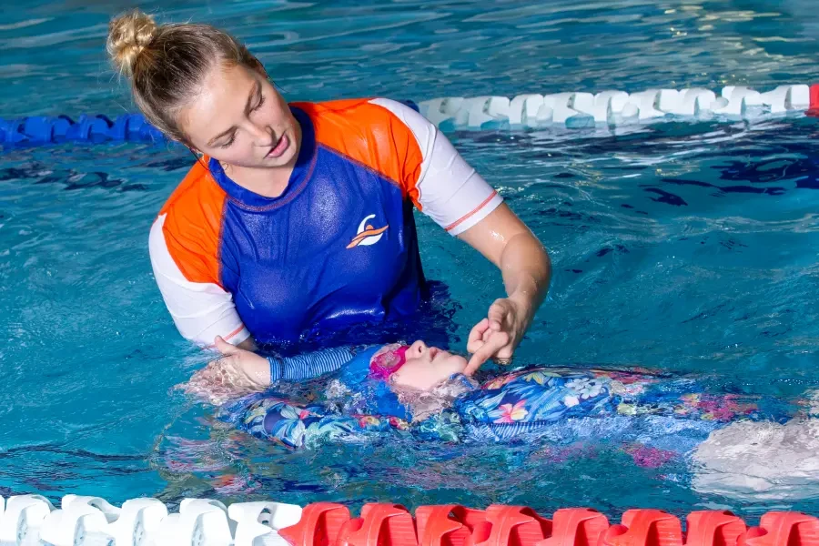 A female swim instructor supports a young girl floating on her back in an indoor pool, gently guiding her body position during a one-on-one lesson.