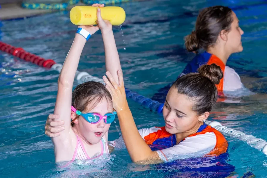 A swim instructor helps a young girl raise a foam pool noodle overhead with both arms during a swimming lesson in an indoor pool, with another instructor and student visible in the background.